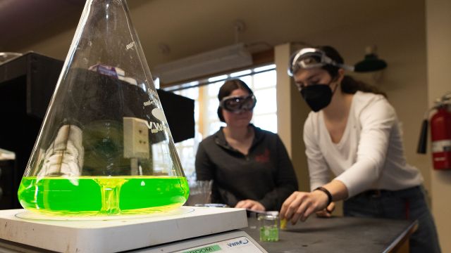 Chemistry Beaker With Yellow-Green Substances With Students Studying In Background