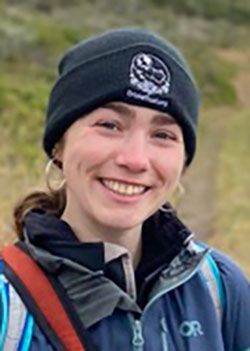 Headshot of Alex Hys wearing a black beanie hat, hoop earrings and a blue jacker.