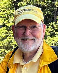 Headshot of Bruce Bishop '68. He is wearing a yellow hat and yellow jacket with a light yellow collared shirt. He wears glasses and has a white beard.