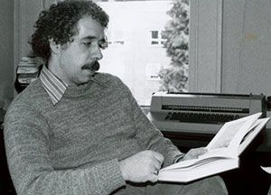 A photo of Mike Steele from 1983. He is sitting in a chair, reading a book.