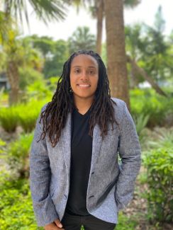 An image of Dr. Rochon outdoors. They are wearing a black shirt, gray blazer, and black pants. 