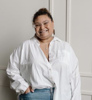 Black woman with curly hair in a bun, wearing glasses, earrings and a white button down shirt smiling at the camera.
