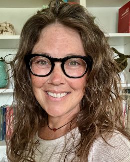 This is a headshot of Cristy Sellers. She is a white woman wearing a beige shirt, black rimmed glasses and she is standing in front of a white bookcase with plants, pottery, and books. She has wavy hair, freckles, and a nose ring. There is a necklace around her neck with a carnelian bead. 