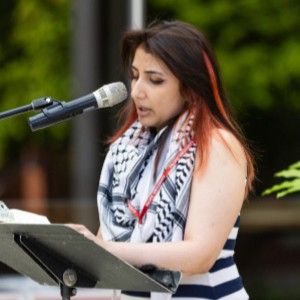 Woman with dark hair with a red streak in it standing at a music stand with a microphone reading poetry on the University Center patio on the Pacific campus in Forest Grove, OR. 