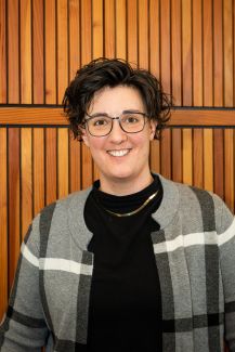 Dr. Mulcahy (a white queer person with black glasses and short curly dark hair, wearing a black turtleneck and grey/blue plaid sweater)) stands in front of a wooden background