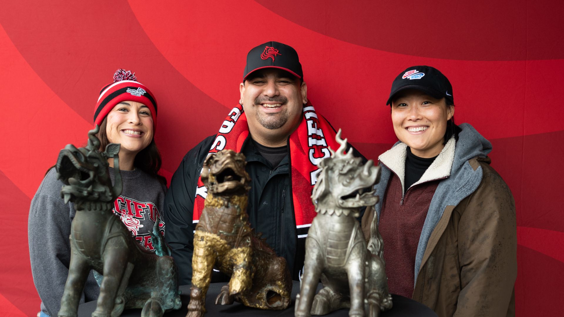 Photo of three people wearing Pacific gear standing behind the three versions of the Boxer statue in front of a red backdrop at Homecoming 2025