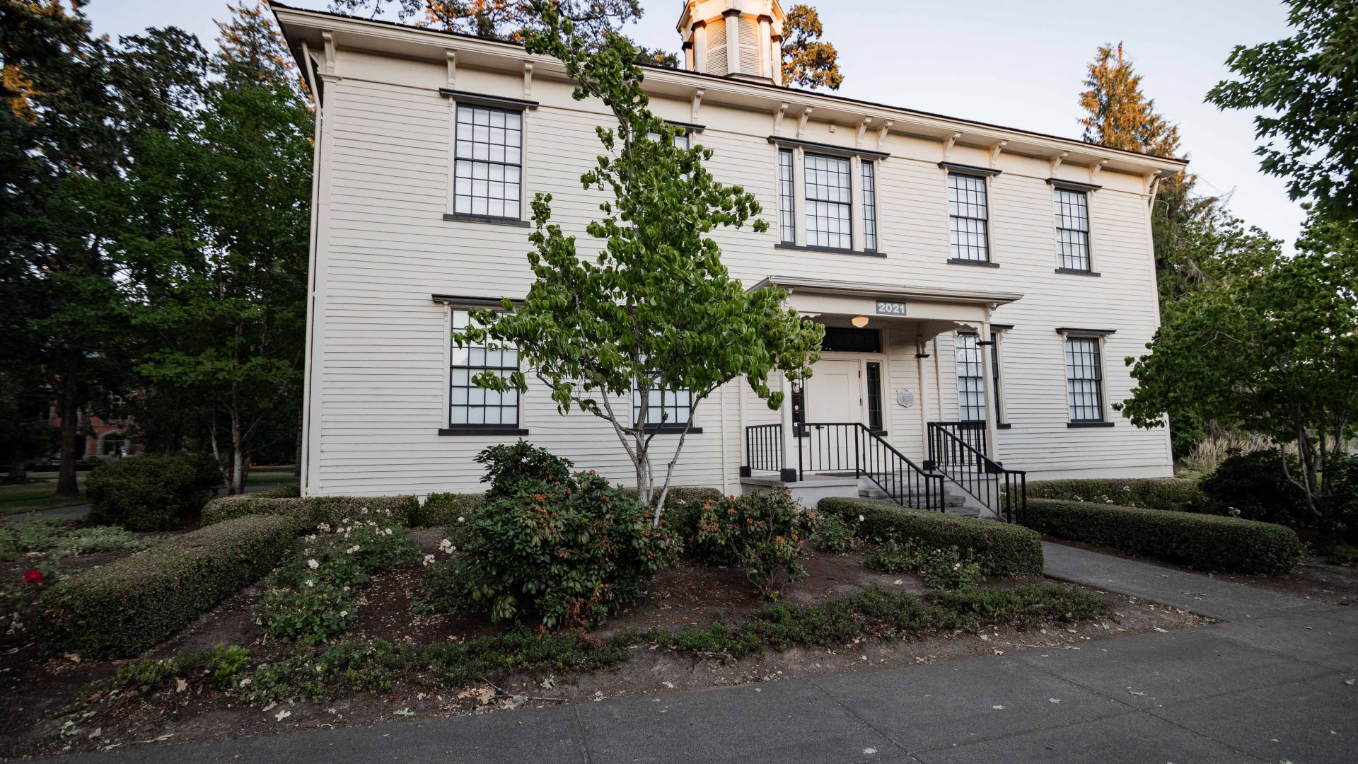 Photo of Old College Hall taken by Kent J Edwards in 2023. It is the front of a clapboard white building made in the classical revival style facing the street. The sun is setting and shines on the belltower and there is a small tree with green leaves to the left of the front door.