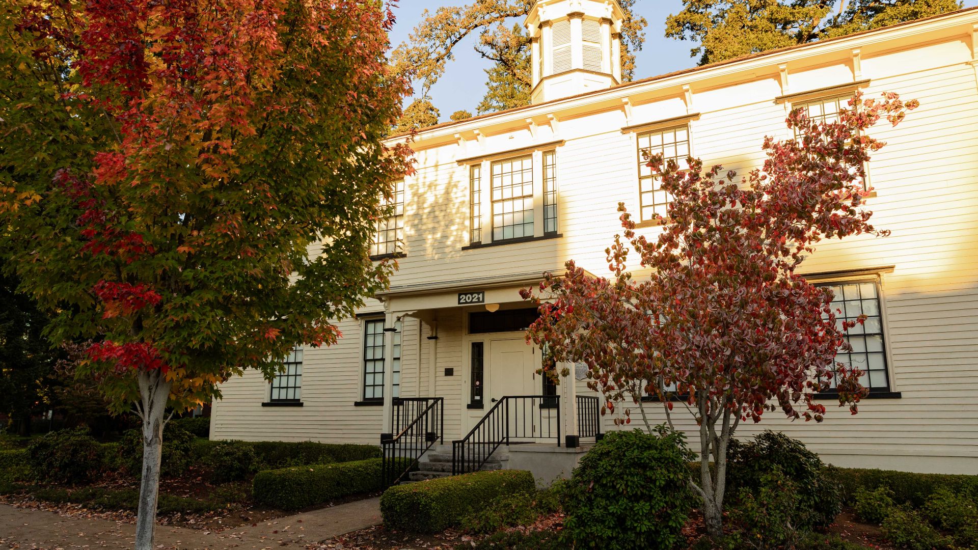 A picture of Old College Hall taken as the sun is setting. The light cuts almost exactly across the center of the building and there are trees with leaves changing from green to orange and red in front.