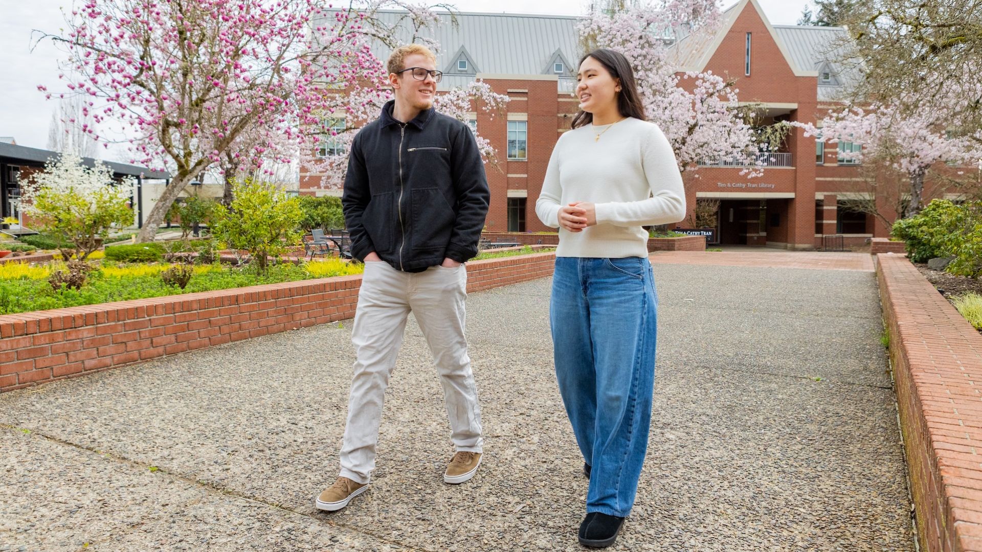 two students walking on the Pacific University campus with a pink flowering tree in the background