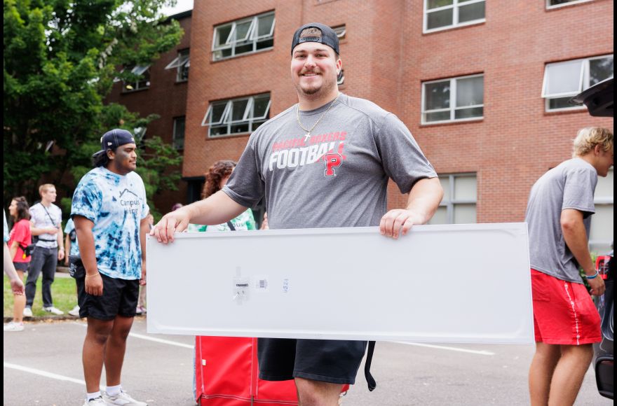 A student wearing a baseball cap and Boxer Football t-shirt carries a mirror into a dorm