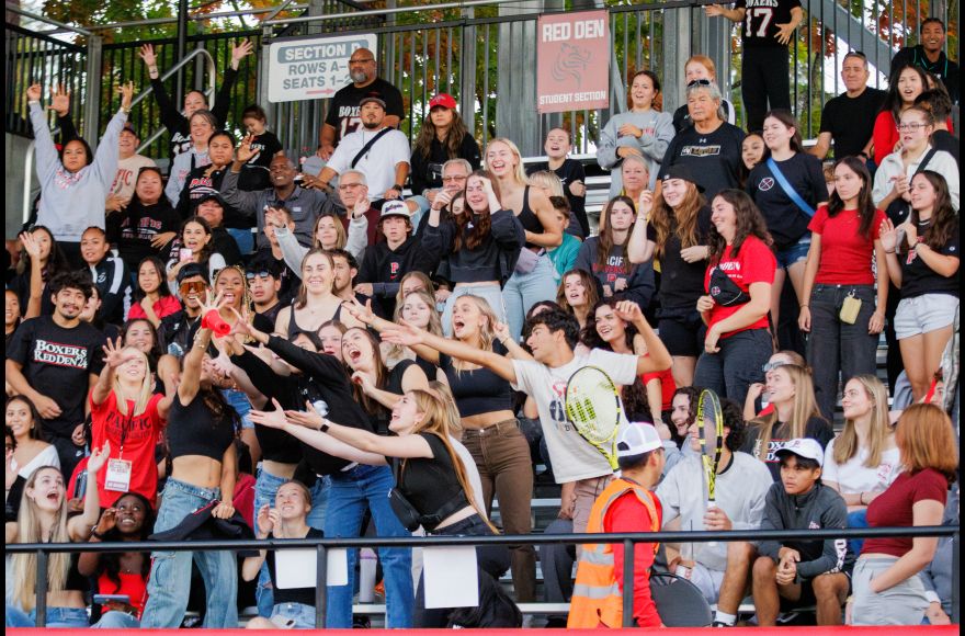 Football fans in the stands stand, cheer, and reach for prizes being tossed into the crowd.