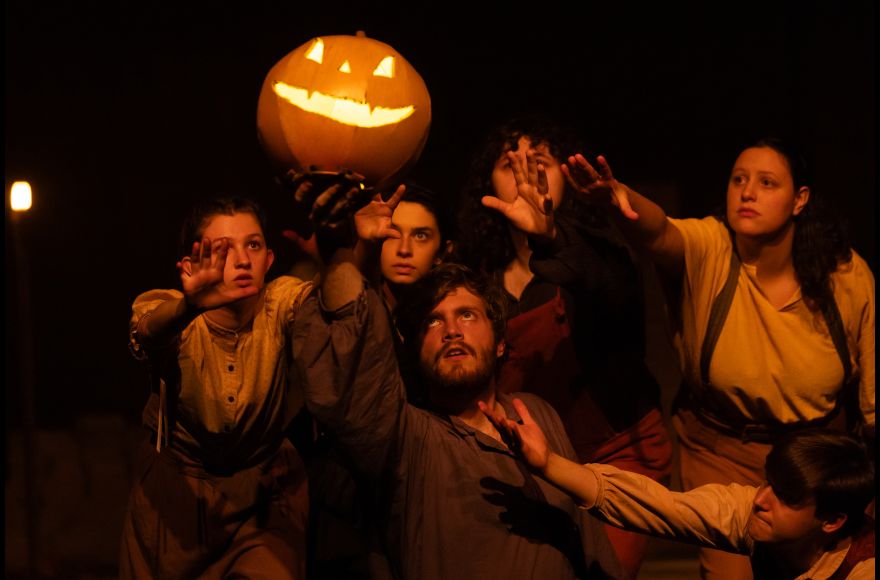 Shadowed students on stage reach for a floating jack-o-lantern during a play